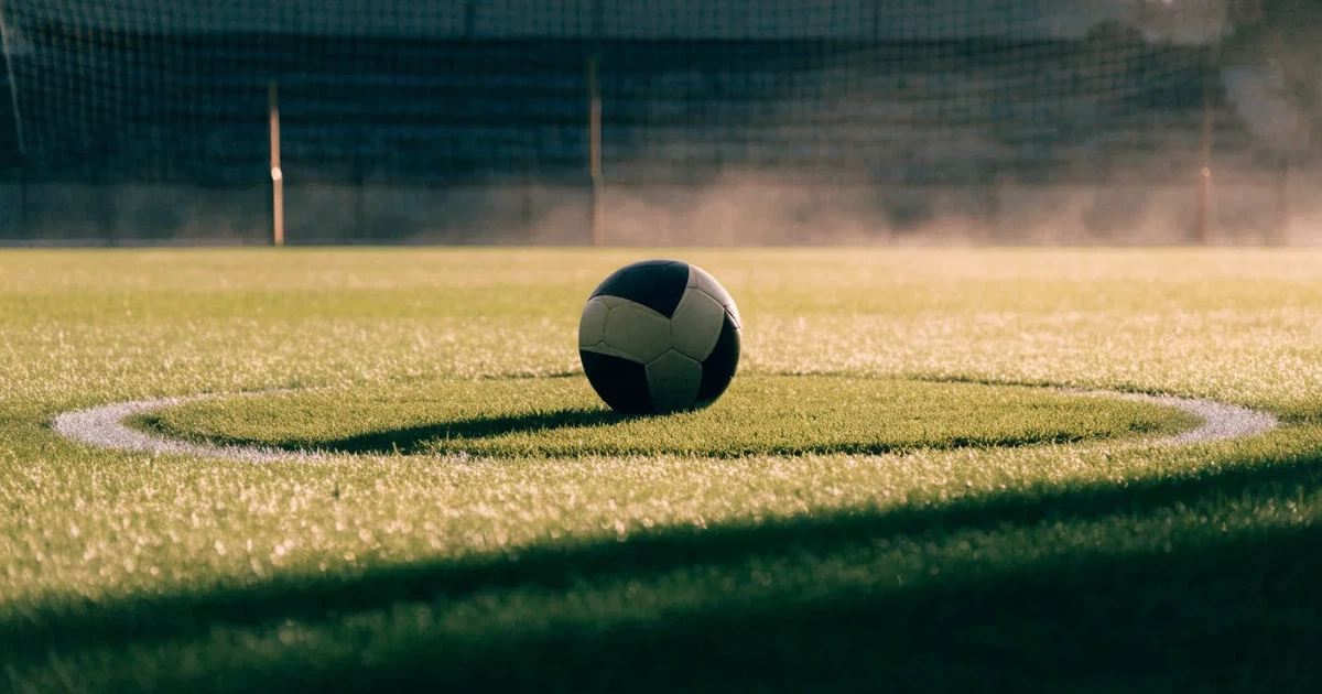 Uma bola de futebol no ponto central de um relvado, à luz da manhã